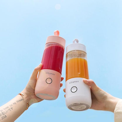 Two hands holding Vitamer rechargeable personal blender bottles with pink and white bases filled with red and orange smoothies